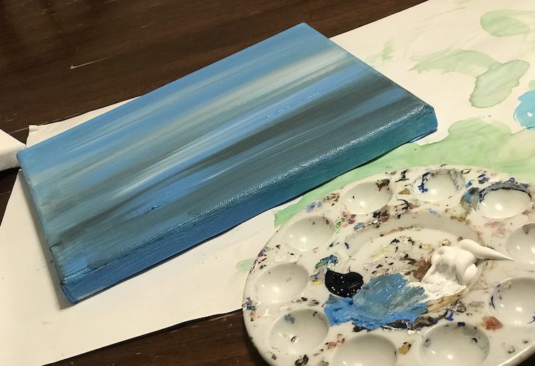 Foreground shows some of a paint palette showing a smudge of blue paint where it used to sit. The black dab and white dollop next to it is only partially used. The finished horizontally streaked blue, white, and black canvas is drying behind the palette. 