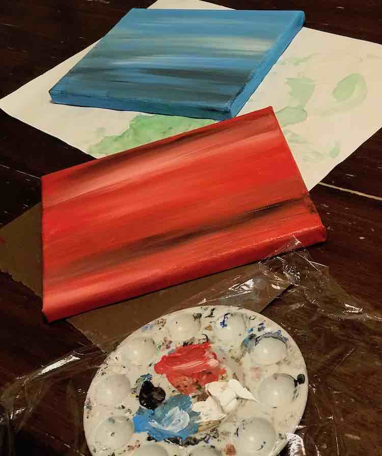 Vertical image with the blue canvas at the top, the red in the middle, and the used paint palette with plastic wrap laid overtop. 