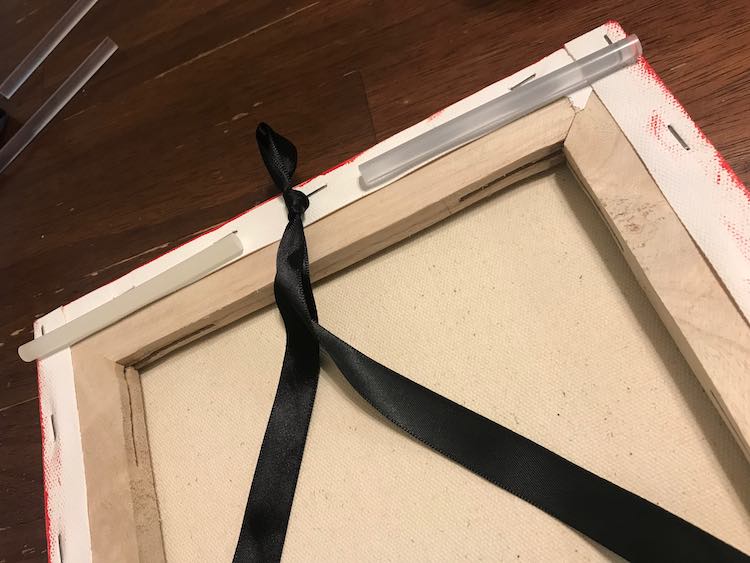 Upper back of the canvas is shown. There's two cold glue stick sitting along the top of the frame each even with the side of the canvas. The ribbon loop is centered between the two glue sticks' inner ends.