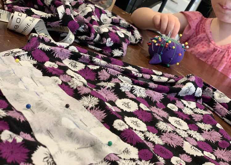 Foreground shows the shirt on the table with the placket pinned in place on it. Since the fabrics are right sides together you can easily see the sketched on line on the placket facing. In the background Zoey is pulling a pin into or out of my purple pin tomato. 