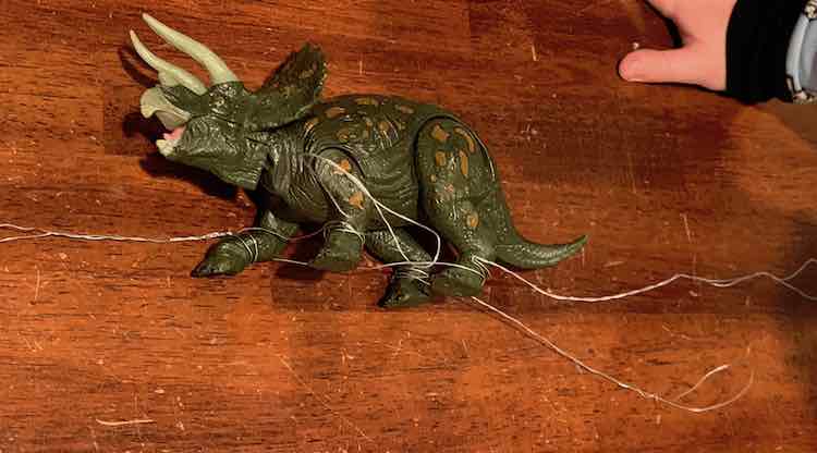 The dinosaur now rest on the table on it's side. All four legs have been wrapped at it's ankles and there is wire sticking out from all four legs. Ada's hand rests in the upper right corner as she looks at what I did to the dinosaur. 