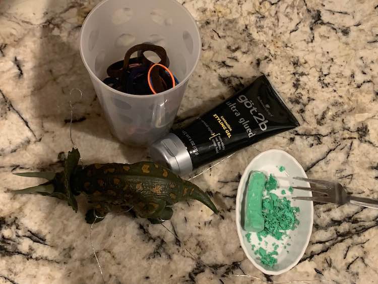 Our counter top with the dinosaur and it's wire framework, the styling gel, a large plastic cup of hair ties, and a dish with partially scraped green sidewalk chalk and a fork leaned over it.