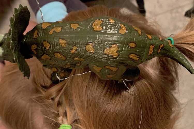 Side view of the triceratops on Ada's head. The front (bottom left in the image) leg is secured by French braiding the wire into place. The bottom of the image shows the lime green hair tie used to keep the braid secure. The second braid is hidden behind the dinosaurs tail but you can see it's hair tie right above the tail. 