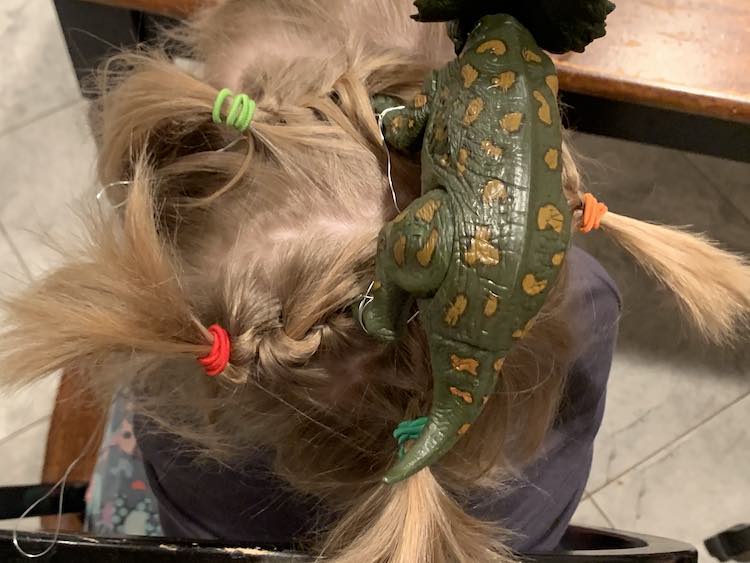 Top view of the dinosaur. You can see all the braids pointing away from the dinosaur. Each one has a different color of hair tie creating the four ponytails: lime green, green, red, and orange.