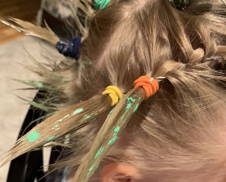 Side view of Ada's hair showing two ponytails in the foreground. Both ponies (orange tie from a braid with a yellow ponytail tie behind) end with a spike of hair pointed away from Ada's head and are dappled with green spots. In the background you can see the back of Ada's short hairs spiked up and a couple other ponytails.