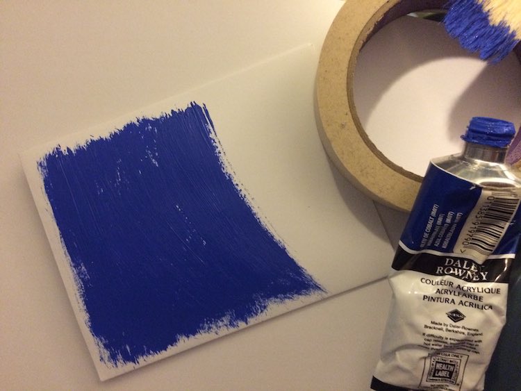 Card on it's side showing the painted sky brushed on quickly. The right of the image shows a roll of masking tape with the blue paint leaned against the bottom and the tip of a paintbrush with blue showing leaned against the top of the tape.