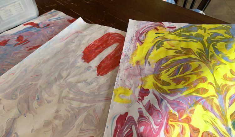 Three sheets of marbled paper laid out on the table. The right one shows red and blue marbling with yellow felt pen in all the white spaces. The middle on shows a lighter purple marbling with a red heart drawn on. The back paper shows purple and blue with red marker sections drawn on.