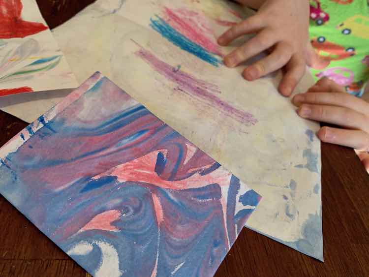 A folded purple and blue card sits at the foreground with red pencil crayon scribbled overtop. The background shows Ada's hands and a slightly marbled paper showing purple, blue, and red pencil crayon marks.