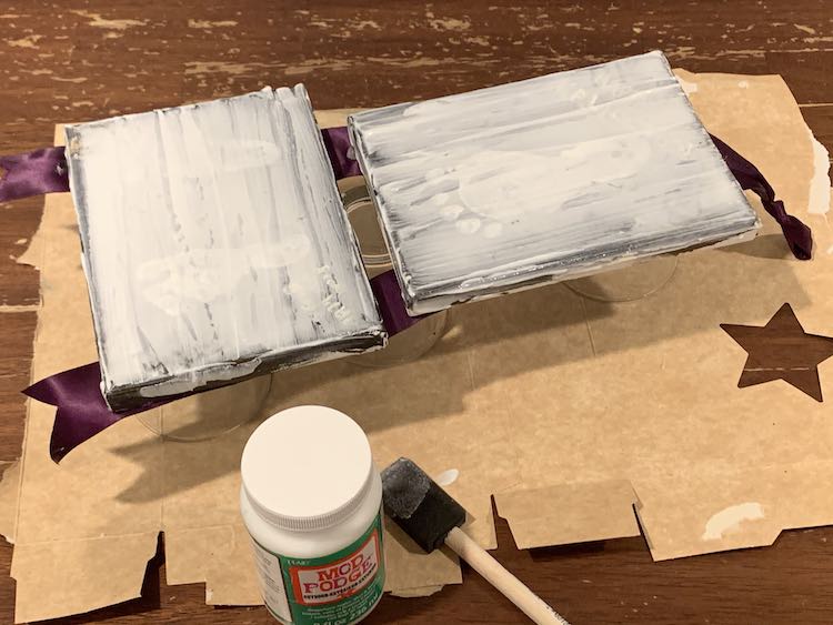 The bottom of the glasses are just peeking out from under the balanced canvases. You can hardly see the image on the canvas as the mod podge is spread on and white right now. Used foam brush and bottle of mod podge are at the bottom of the photo. 