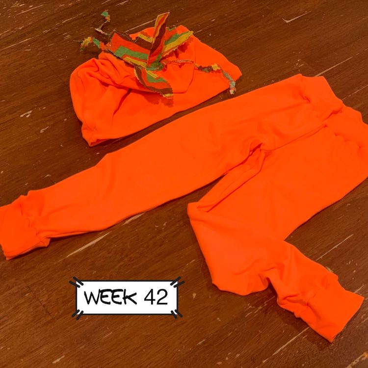 A neon orange beanie with striped leaves laying on a table next to neon orange leggings. The leaves are striped brown, orange, green, and yellow.