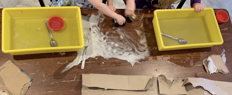 The girls each have a yellow bin full of plain water in front of them. They still have a red plastic cupcake liner and spoon from the earlier shaving cream bin. The space on the table between them is a mix of shaving cream tinted water and they're running plastic toy monster trucks through it. 