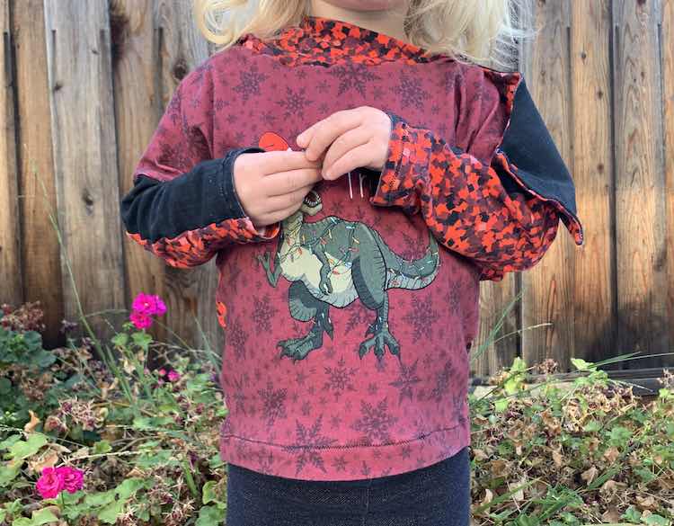 Closeup of the front of Zoey's hoodie with her arms up in front of the "Tree Rex" showing her sleeves and with the flap showing over her arm from the lowered hood hanging behind her.
