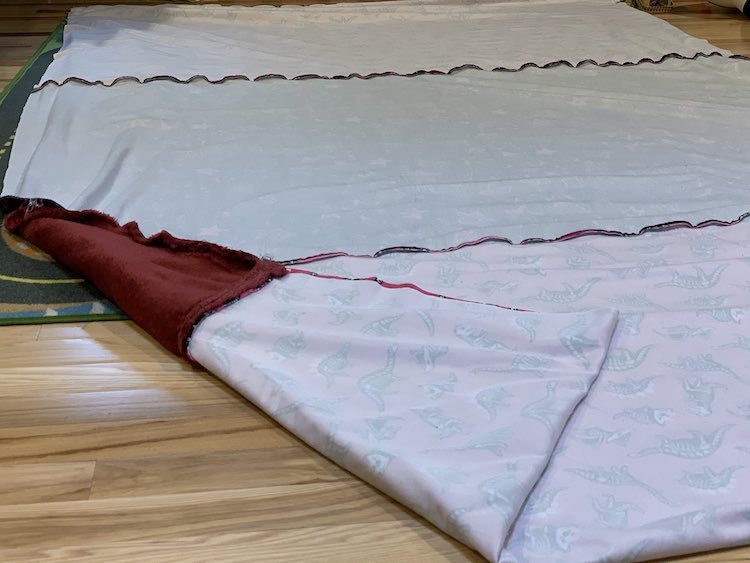 The long blanket has been folded in half, right sides together, so the new center was found and the wrong side is facing up. The folded dinosaur fabric is at the bottom of the picture where the left corner is folded over to show the reverse side and the join between the dinosaur and throw. 