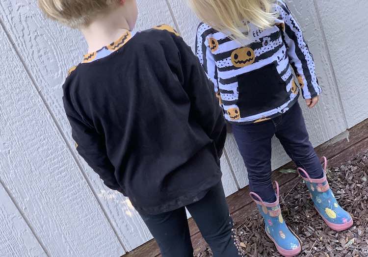 Ada is slightly closer to the camera facing Zoey so Ada's back is to you and Zoey's front is facing you. You can see the back of Ada's top showing the pumpkins on the back of her neckband. Zoey's hair is obscuring the top of her top. 