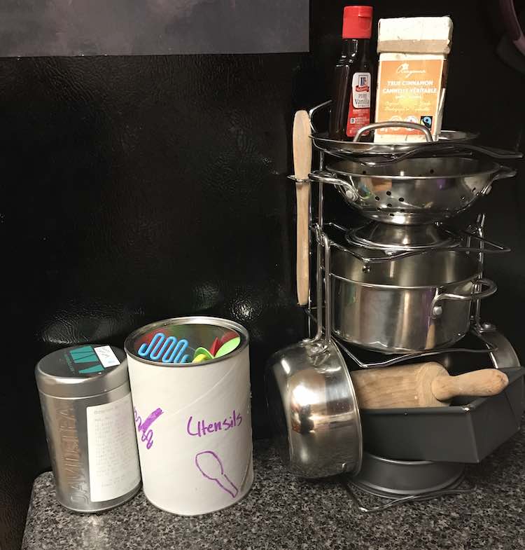Front view showing the Melissa and Doug stand with pots on it, some empty cinnamon and vanilla container on top, and child’s baking set underneath. To the left is a white canister with plastic utensils inside and then beside that is an empty David’s tea canister to play with. 