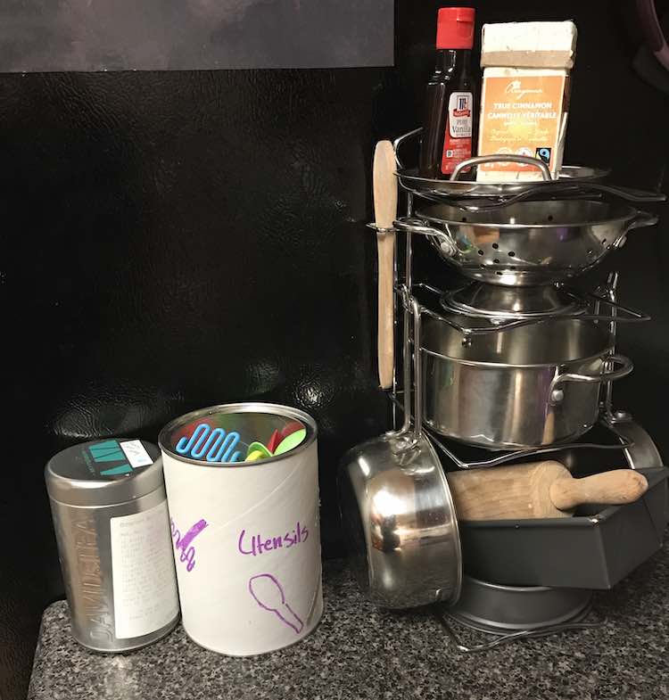 Front view showing the Melissa and Doug stand with pots on it, some empty cinnamon and vanilla container on top, and child’s baking set underneath. To the left is a white canister with plastic utensils inside and then beside that is an empty David’s tea canister to play with.
