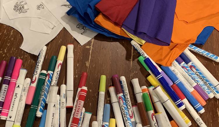 The image shows varying hues of fabric markers spread across the table and along the bottom of the image. The upper half of the image shows the stacked bodice pieces, to the left, and the pile of cut, presewn, skirt pieces to the right and overlapping the pattern pieces and some of the markers. 