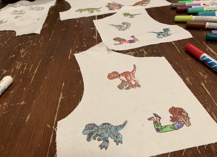 This image shows all three of Ada's bodice pattern pieces. The closest piece, in the foreground, shows a LEGO® Friend laying down and two dinosaurs. In the background you can vaguely see the other half back showing and another LEGO® friend and three dinosaurs. In the very back of the image you can see the bottom of the front bodice piece with the group of Friends and two dinosaurs facing away.