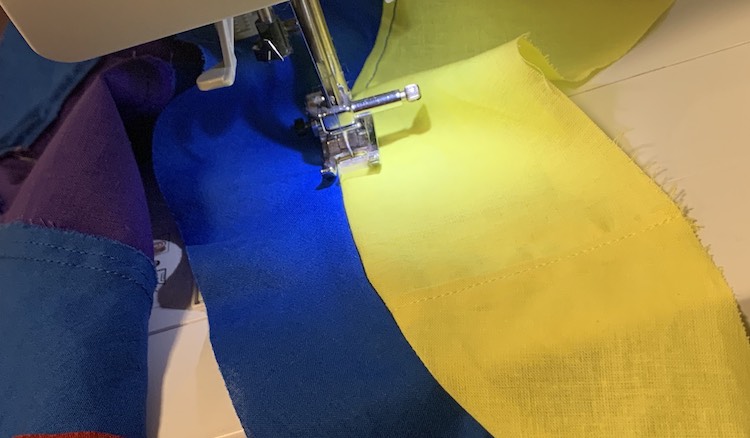 My sewing machine is shown while it top stitches the seam down between the blue and yellow swirl. Off to the left you can see the underside of the topstitched seam between the blue and purple swirls. 