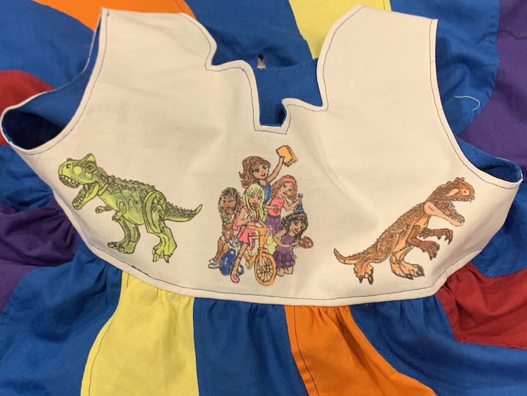 Closeup of the front of the bodice showing the group of LEGO® Friends surrounded by two, looking away, Tyrannosaurus Rex dinosaurs. Surrounding the bodice are the skirt's swirls going away from the bodice and the visible blue liner from the inside of the bodice.