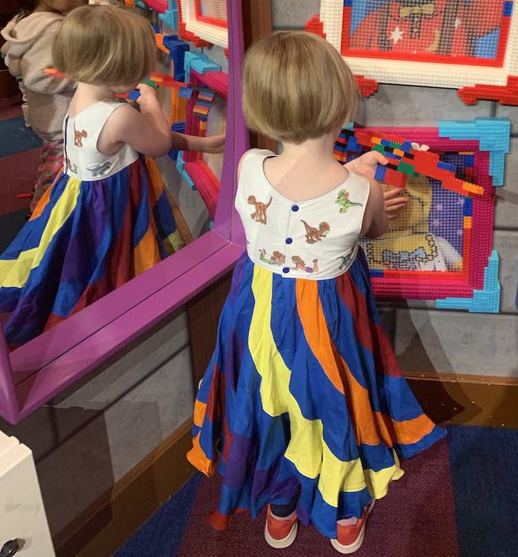 The back of Ada is shown as she holds her LEGO® structure and touches a LEGO® picture brick frame while standing beside a mirror.