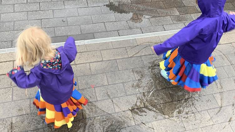 Photo is taken from above looking down at the girls' backs while they stand in puddles wearing rain jackets. 