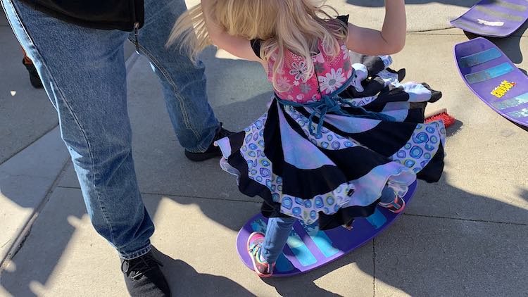 Photo taken looking down at Zoey on a balance board. She's holding her dad's hand, on photo, and getting twirled.