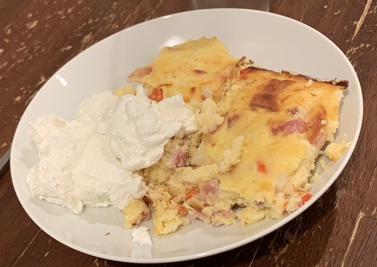 A pancake shaped piece of casserole on a white plate with a dollop of yogurt to the side.