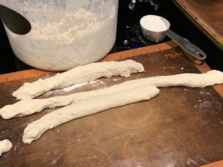 Image shows the pail of bread dough near the top with a silpat below it. On the mat are three bread dough 'snakes' (cylinders). The middle one is the longest and half as skinny as the others. 