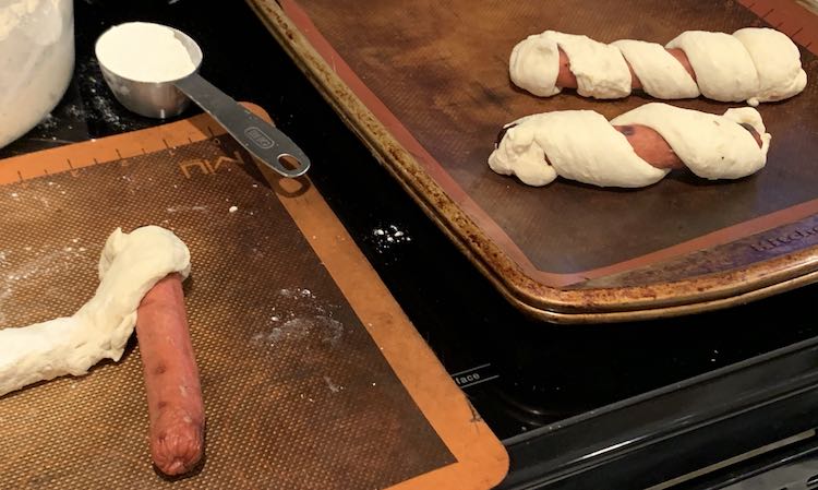 Shows a silpat with a partially wrapped hot dog weiner (showing how you start it off). The background shows a cookie sheet with two mummified hot dog weiners.