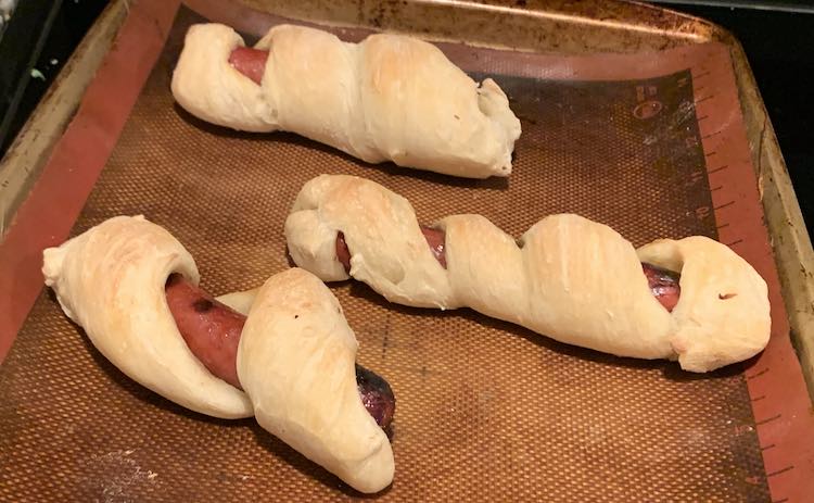 Closeup of the end of the cookie sheet lined with a silpat and showing three cooked mummified hot dogs.