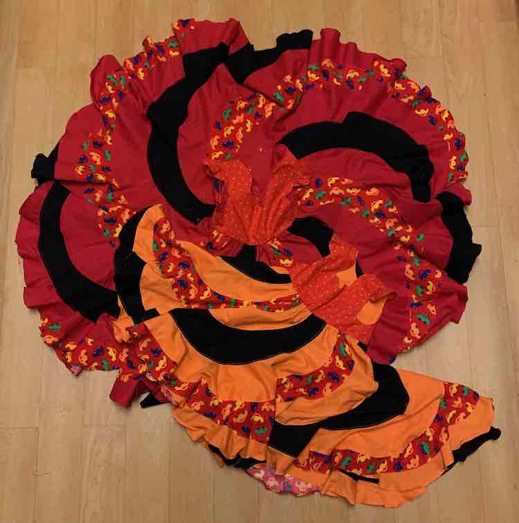 The two dresses laid out on the floor with the photo take above looking down. The larger, red, dress has it's skirt completely open so it's a circle with the bodice in the center. The smaller, orange, dress is laid out at an angle at the bottom. 