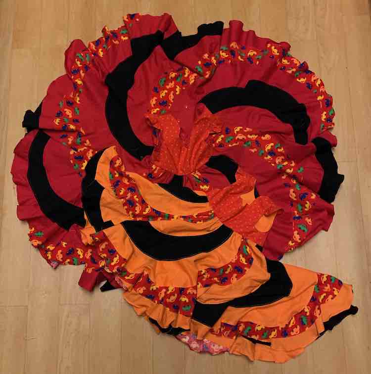 The two dresses laid out on the floor with the photo take above looking down. The larger, red, dress has it's skirt completely open so it's a circle with the bodice in the center. The smaller, orange, dress is laid out at an angle at the bottom.