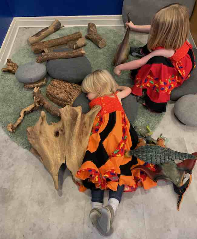 Zoey lays on her tummy on the green grass carpet surrounded by toy dinosaurs and driftwood. Her upper body rests on a 'stone' pillow. Ada sits on her own 'stone' pillow next to her while playing with the brontosaurus. 