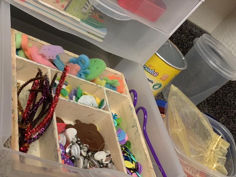 Closeup of the bottom drawer showing a repurposed Melissa and Doug stamp container holding a pipe cleaner tree, buttons, crazy straw, felt circles with holes, mini shaped sponges, beads, and a dollar tree balloon weight. To the side is some empty containers and small fabric bag to play with. 