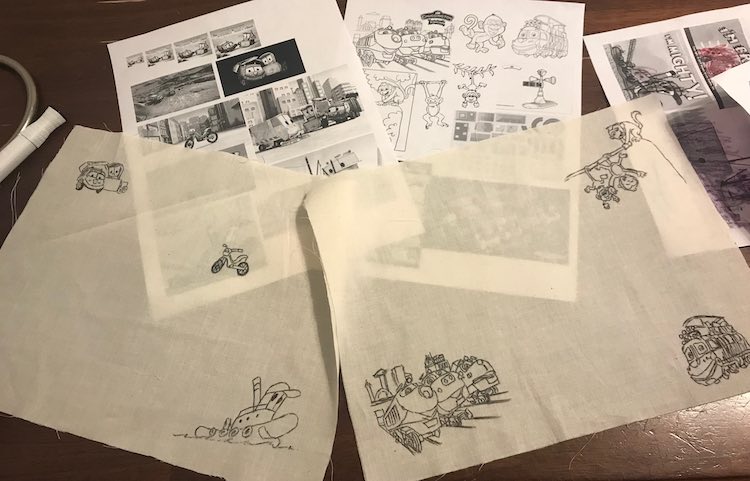 Two rectangles of fabric slightly overlapping showing the characters in black fine-tipped fabric markers. The coloring pages they were traced from are slightly above and behind the fabric. 
