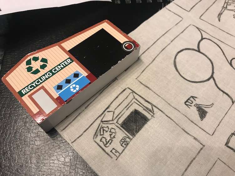 Closeup of the upper left side of the fabric showing a simplified recycling center sketched out. Above it is the 2D wooden Melissa and Doug recycling center toy that I used as a model to sketch it out.