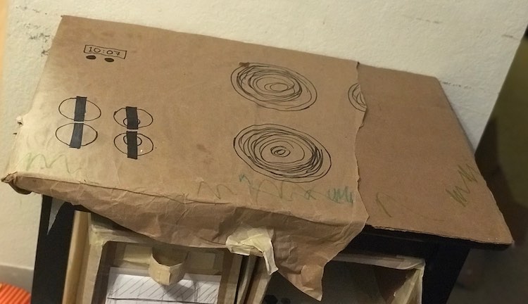 Angled top view of the kitchen set before removing the top. Two thirds of it is covered in brown paper showing the four dials for the burners, and timer, and two of the burners. The right side, with the remaining two burners, had been ripped off a while back and shows a sheet of cardboard beneath it. There's green scribbles across the front of the paper.