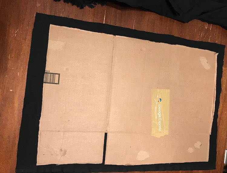 Photo is an overhead view of the kitchen table showing a rectangle of black fabric peaking out from under the piece of cardboard. At the top of the image you see more black from the main piece of fabric that the first piece came from.