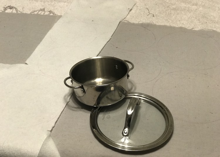 Image show the grey flannel and some of the white interfacing laid out. On top of them is a small actual kitchen pot lid and a smaller play pot. Behind the items you can see black outline of circles where the items have already been traced out.