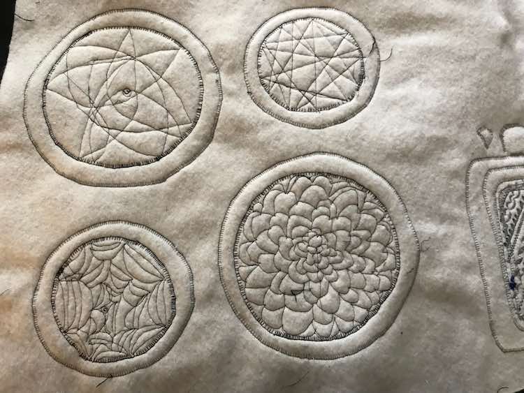 Closeup of the underside of the kitchen set. The black bobbin thread shows up against the white interfacing letting you easily spot the drip trays (circles) and the burners (free motion quilting patterns). The left top burner is a range of curves ended with points causing a flower-like star. The small top right burner shows straight lines bisecting the circle although there's too many to be considered a star. The small circle at the bottom left almost looks like a spider web while the large circle to the right looks like a many petaled flower akin to a dahlia.
