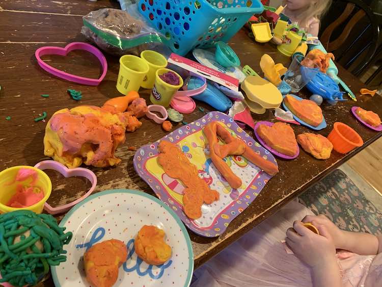 Playdough cookies plated on many different plastic plates and a tea tray. The plates, playdough, and tools are spread across the table while both kids sit at the table creating. 