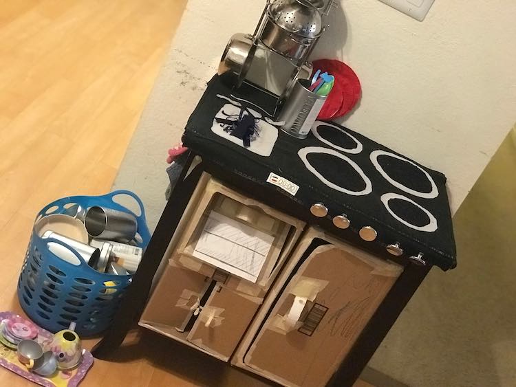 Front view showing the cardboard appliances within the reclaimed nightstand. The new stovetop is visible with it's already drooping knobs and the pots and pans stacked neatly. 