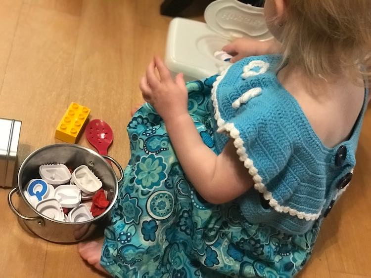 Zoey sitting on the floor with a pot of plastic letters beside her. She's currently inserting a letter into an empty plastic Huggies wipe container.