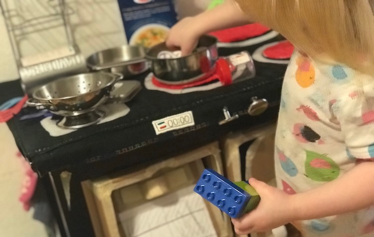 Front view of the kitchen set with a strainer sitting in the kitchen sink and a pot on a turned on burner. Ada is currently using her hands to add DUPLO® to the pot.