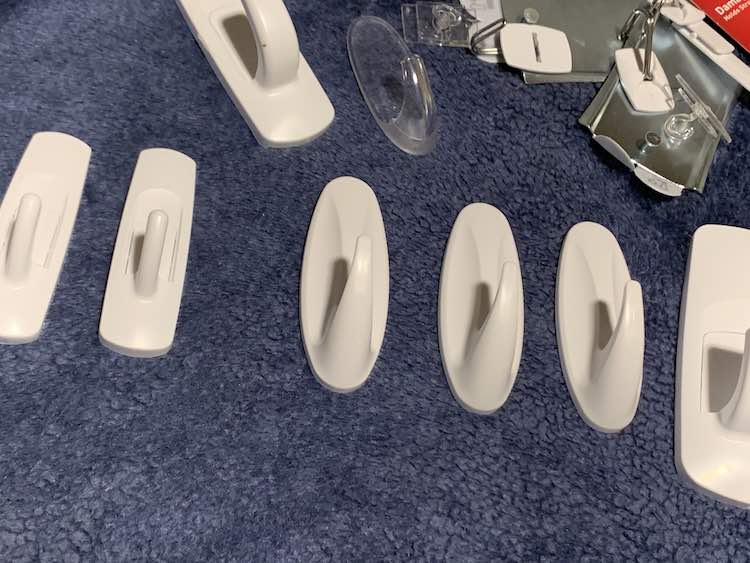 Blue blanket with the command hooks spread out on them. On the far left are two medium-sized rectangular command hooks followed by three medium oval hooks, and then a large rectangular hook. Above them are a random assortment of other command hooks mostly too small.