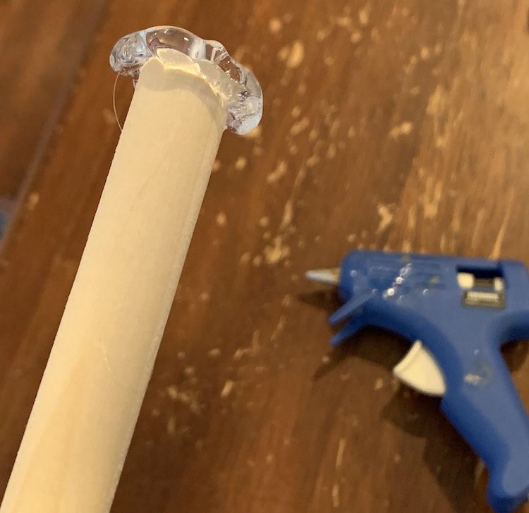 Closeup of the dowel end with transparent glue attached to the end of it. I the background you can see the slightly blurry blue glue gun laying on the kitchen table. 