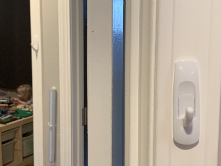 The picture is taken in the living room doorway showing the right side of the doorframe with the rectangular command hook. Through the doorway you can see the wall and the bedroom doorway with another command hook. On the far left of the image you can see a blurry Zoey playing in her bedroom.