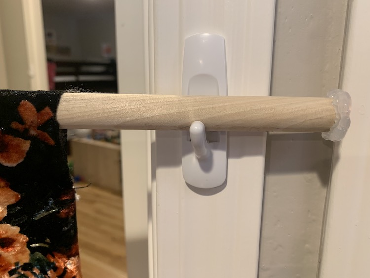 Center of the image shows a command hook attached to a white door frame. On the left you can see the edge of the curtain on the dowel and on the right side you see the glue covered tip of the dowel. 