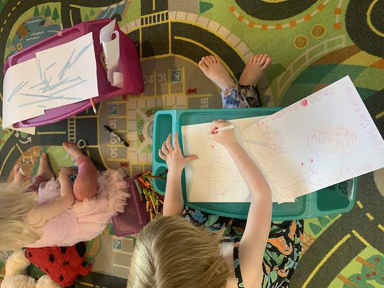 Image shows an overview of Ada and Zoey sitting on the car mat drawing on lap desks. Zoey is using decorative washi tape to attach to pieces of paper together artistically while Ada, to the right, follows the video to draw the pigeon from Mo Willems' book. 